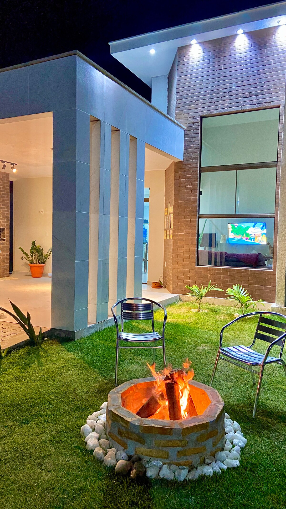An outdoor seating area features two metal chairs arranged around a stone fire pit, where warm flames dance. Lush green grass surrounds the area, and the structure’s modern architecture provides a backdrop with large windows that reflect soft indoor lighting.