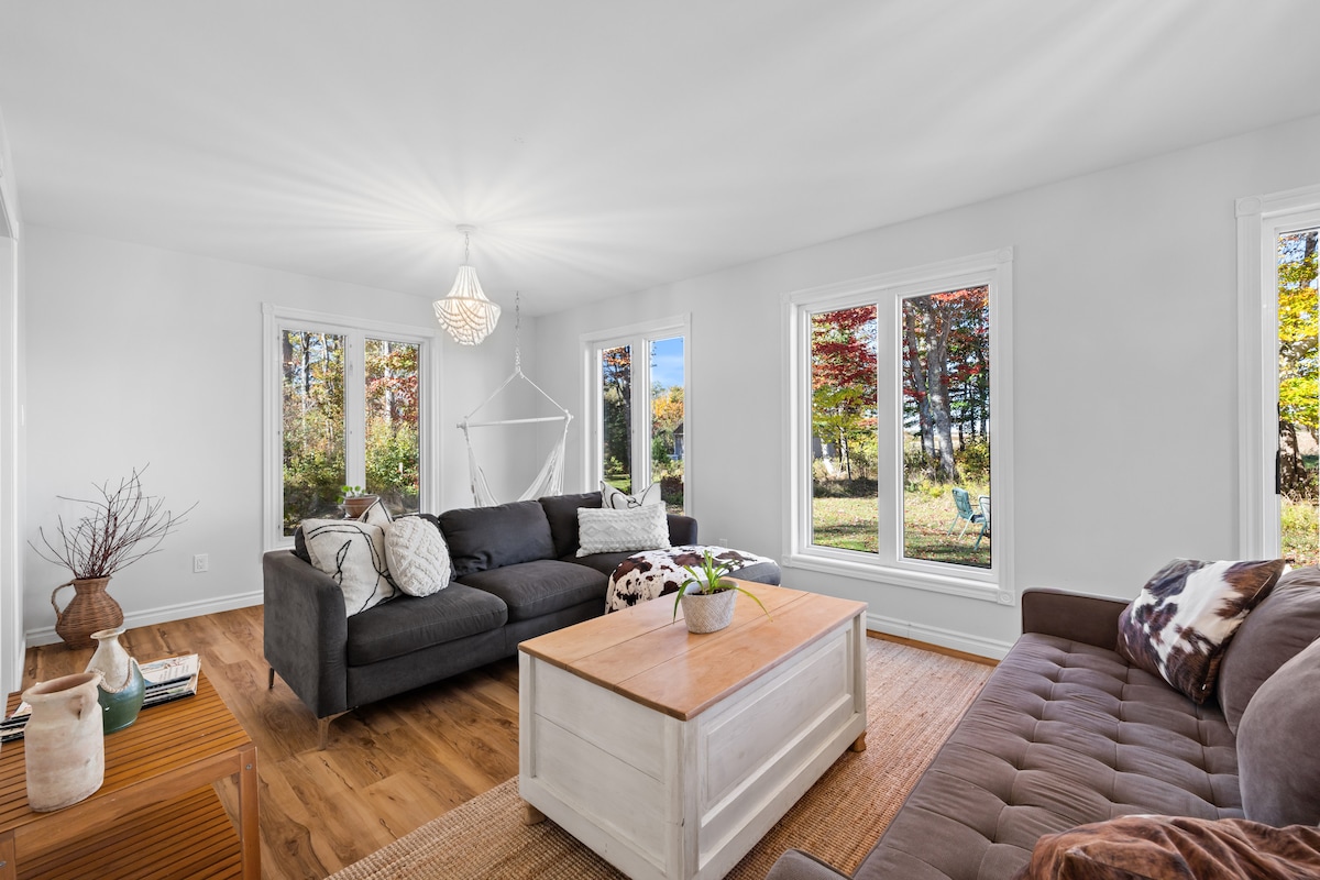 A bright living room is filled with natural light, featuring two large couches and a wooden coffee table at center. Large windows reveal a view of colorful trees outside. A simple light fixture hangs from the ceiling, enhancing the spacious layout.