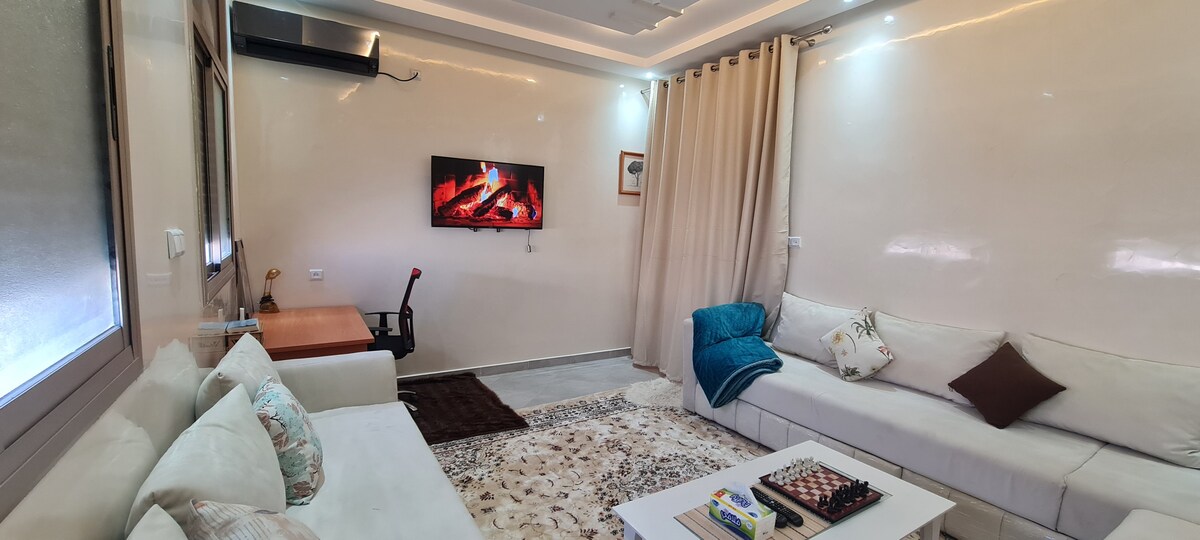 A well-lit living area features a comfortable seating arrangement with two sofas and a coffee table. A wall-mounted TV displays a virtual fireplace ambiance. Soft neutral tones are complemented by decorative cushions and a plush area rug, creating a cozy and welcoming environment.