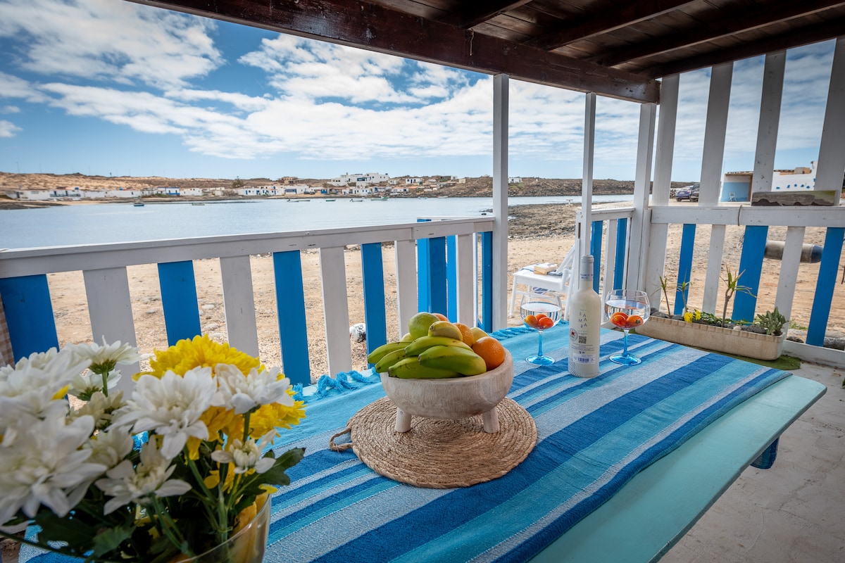A table is set on a shaded terrace, adorned with a woven placemat holding fresh fruit. Clear glasses containing a drink rest on the table, complemented by a white floral arrangement. The view includes a serene coastal landscape under a partly cloudy sky.