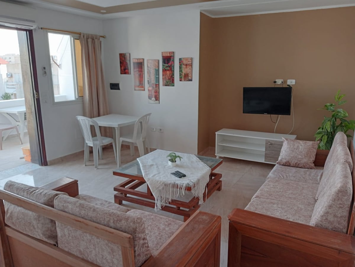 A bright and airy living area is showcased, featuring wooden furniture with comfortable seating. A small dining table with plastic chairs can be seen beside a wall-mounted television. Decorative wall art adds color, while sunlight filters through the nearby window.