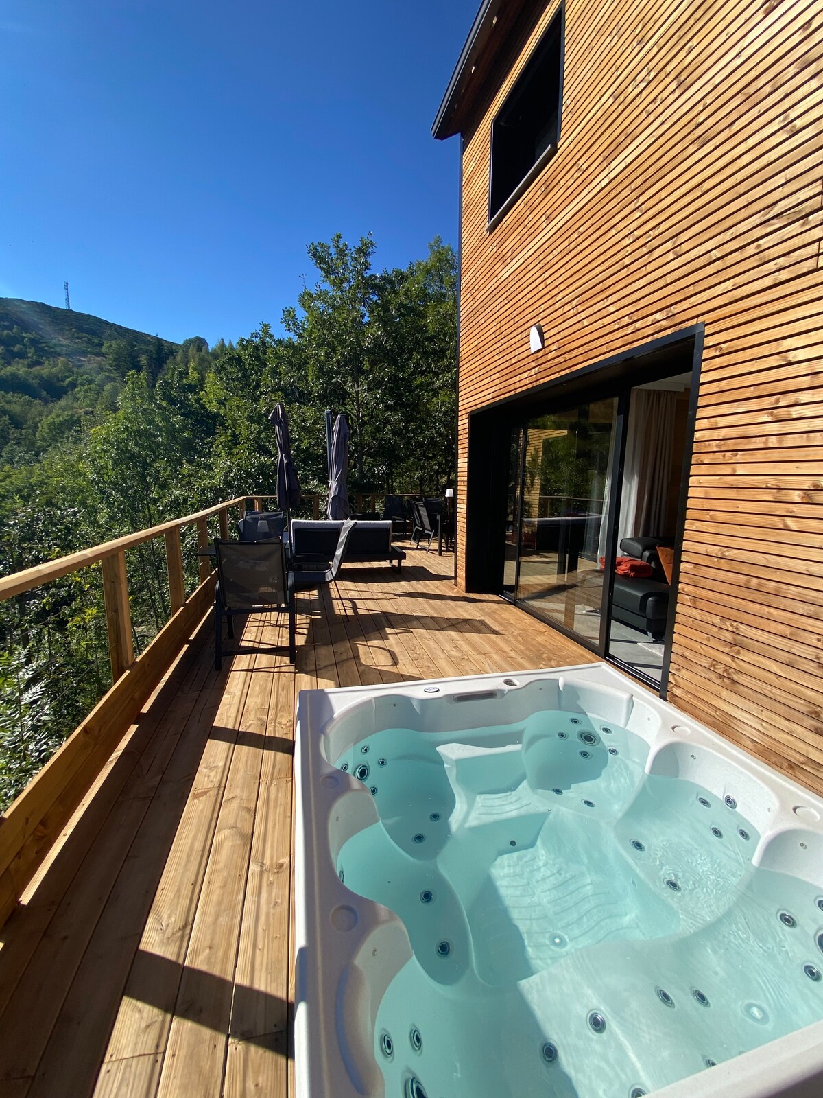 A wooden terrace is presented with a hot tub filled with clear water, surrounded by lush greenery. Outdoor seating, including two umbrellas, offers a relaxing space to enjoy the mountain views in a contemporary setting. Large glass doors invite natural light into the interior.