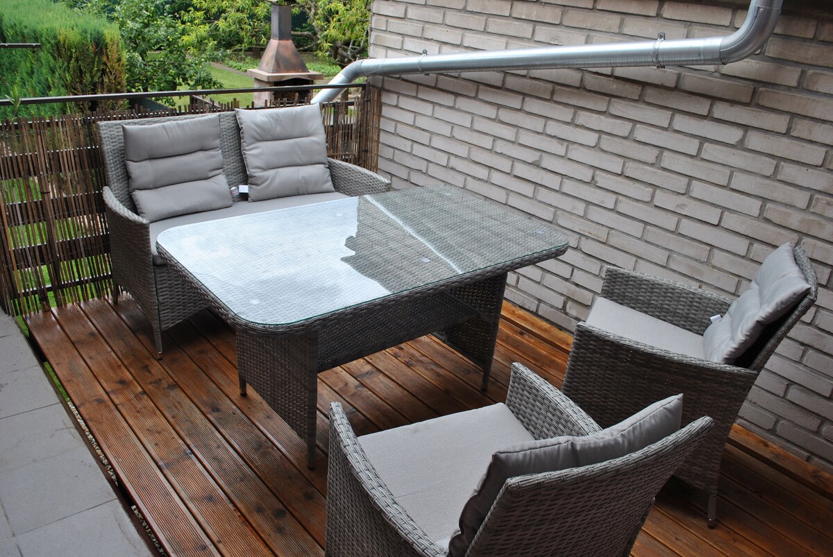 A covered outdoor seating area features a glass-topped table surrounded by four grey wicker chairs with cushions. The wooden deck is bordered by bamboo fencing, and greenery is visible in the background, contributing to a calm environment.
