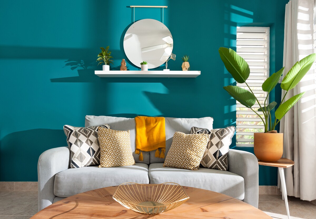 A cozy living area is highlighted by a soft grey sofa adorned with decorative cushions in various patterns. A round wooden coffee table displays a unique bowl. A large mirror is mounted above a shelf with small plants, and a potted plant adds a touch of greenery to the space.