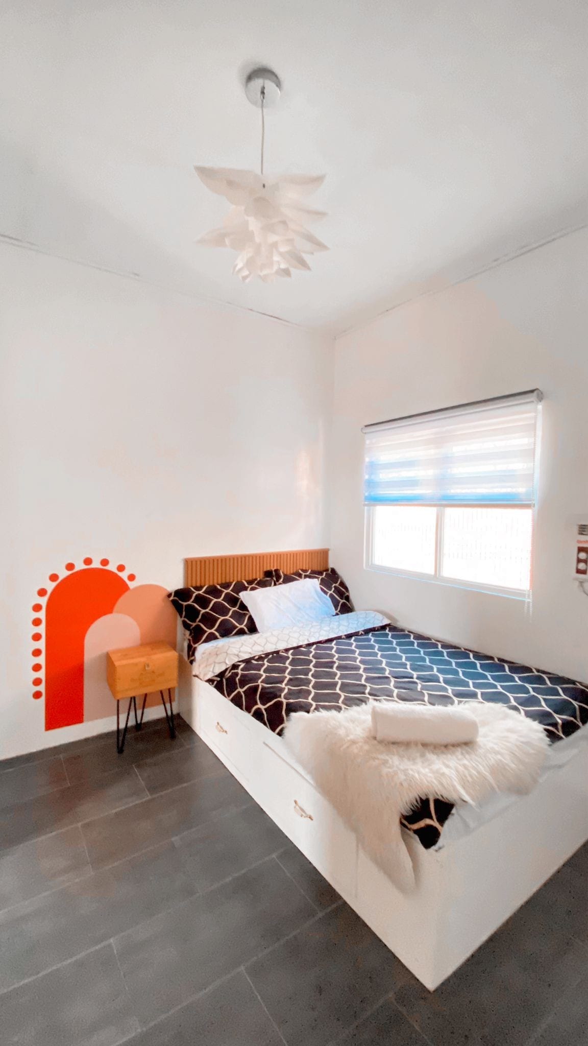 A cozy bedroom features a double bed adorned with a patterned bedspread and a decorative throw. A minimalist chair sits beside the bed. Light enters through a window with layered shades, while a modern light fixture hangs from the ceiling, complementing the white walls.
