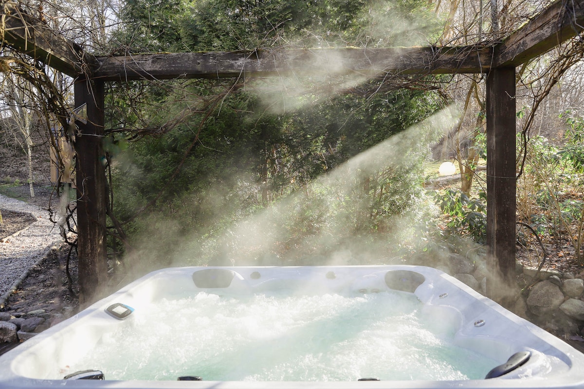 A private outdoor jacuzzi is surrounded by lush greenery, with steam rising gently from the surface of the bubbling water. Sunlight filters through the trees, creating a serene and inviting atmosphere for relaxation.