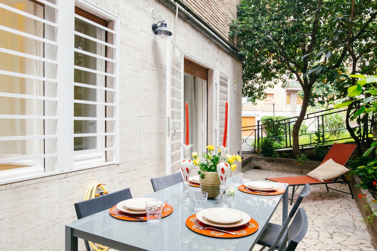An outdoor dining area features a glass table set for four, complete with colorful plates and glasses. Vibrant flowers are displayed in centerpieces, and a comfortable lounge chair is positioned nearby. The space is surrounded by greenery and is accessible through large glass doors.