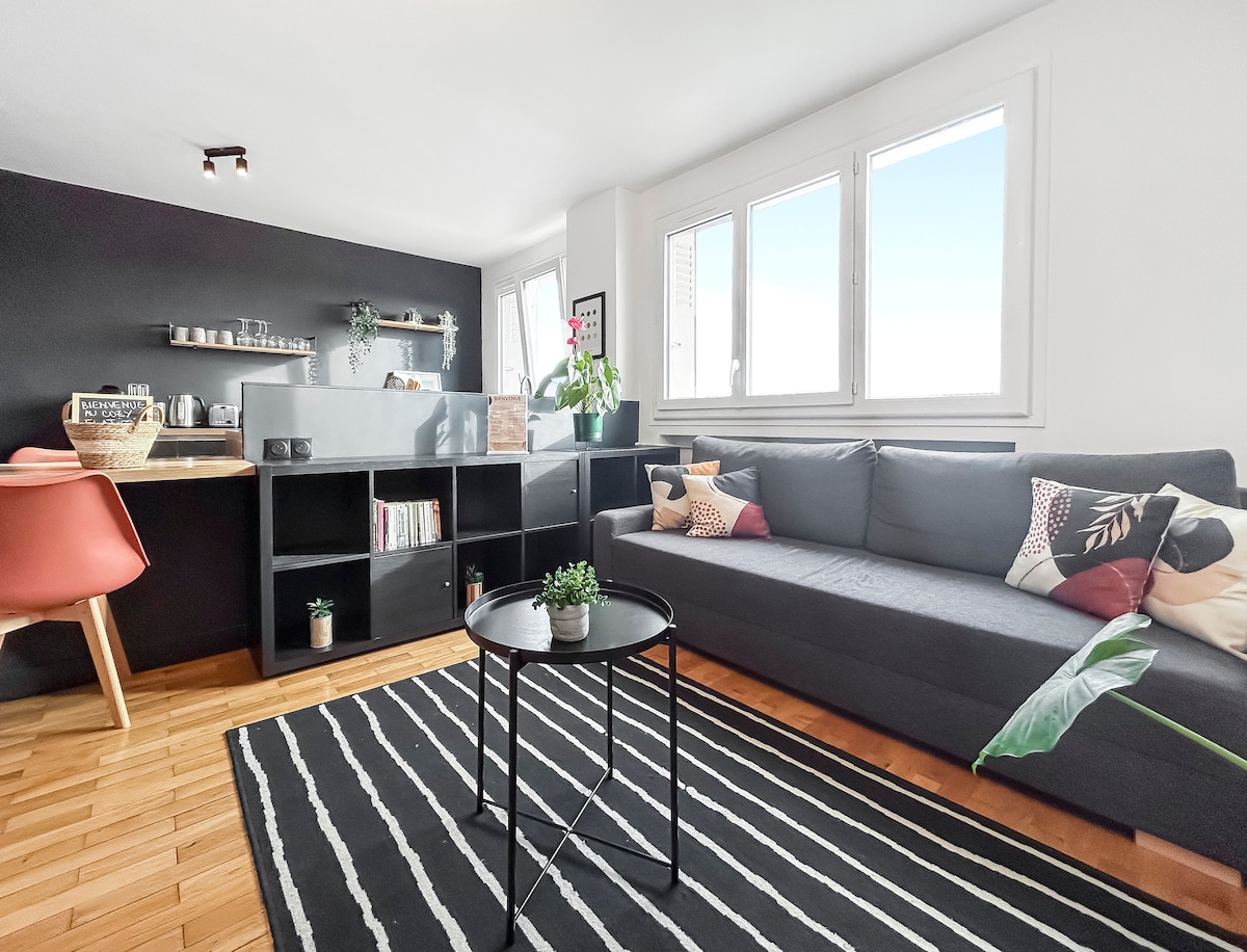 A light-filled living area features a modern grey sectional sofa adorned with decorative cushions. A circular black coffee table rests on a striped rug, while a sleek open kitchen with wooden chairs is visible in the background, illuminated by natural light from surrounding windows.