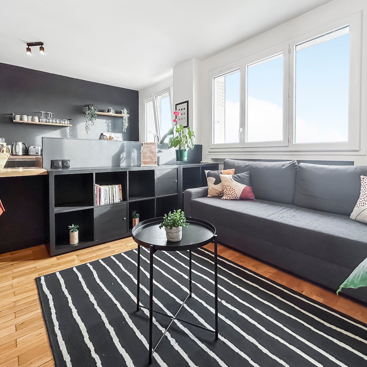 A modern living area features a comfortable gray sofa and a coffee table atop a stylish striped rug. Large windows provide ample natural light, showcasing a minimalist design with shelving and greenery accented throughout the space.