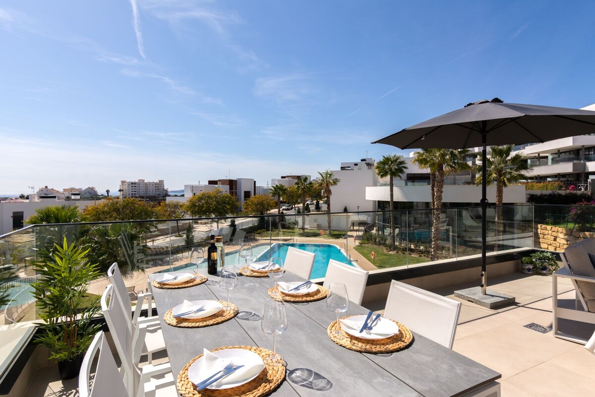 A dining table is set for six on a spacious terrace, with plates, glasses, and decorative linens arranged neatly. The background features a pool area surrounded by palm trees and well-kept landscaping, under a clear blue sky.