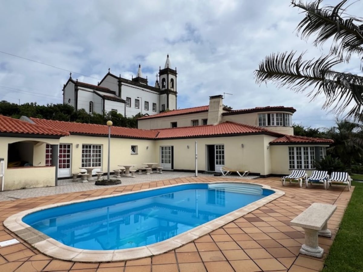 An outdoor swimming pool is framed by a spacious patio surrounded by lounge chairs. The villa's two-story structure is visible in the background, featuring a traditional design with red-tiled roofs and multiple glass doors. Lush greenery adds to the serene setting.