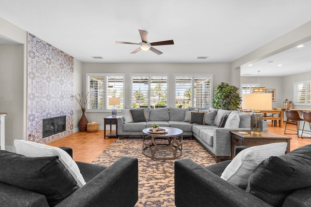 A spacious living area features a large sectional sofa surrounded by accent chairs. Natural light streams through multiple windows, illuminating the room's neutral tones. A patterned area rug adds texture, and a decorative wall with intricate designs serves as a focal point. A ceiling fan offers comfort.