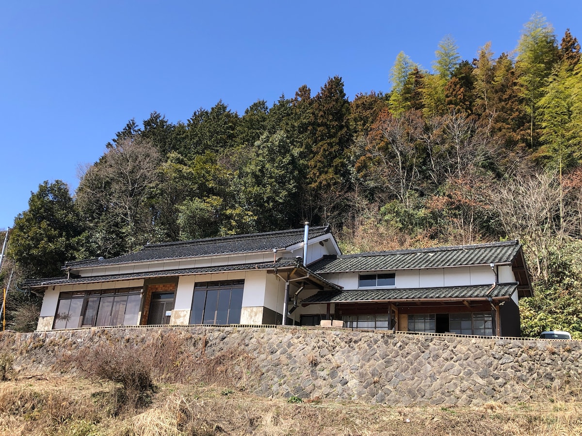 A spacious home is positioned against a backdrop of lush green hills. Large windows allow for ample natural light, showcasing the inviting exterior with traditional roofing. The surrounding landscape includes trees and foliage, contributing to a sense of tranquility.