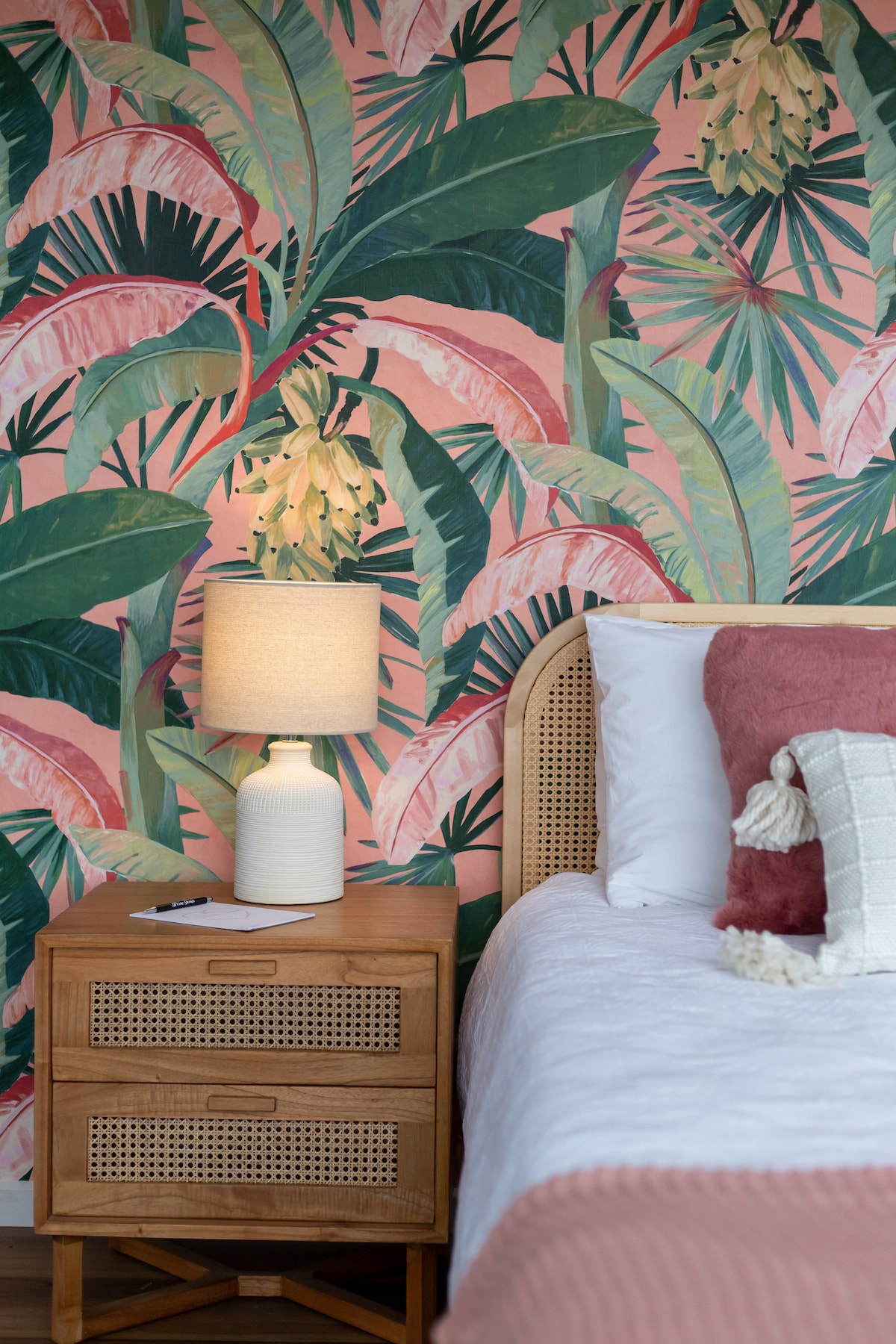 A vibrant wall adorned with tropical foliage creates a lively backdrop in the sleeping area. A textured beige lamp sits atop a wooden nightstand, complementing the soft pink tones in the bedding and decorative pillows.