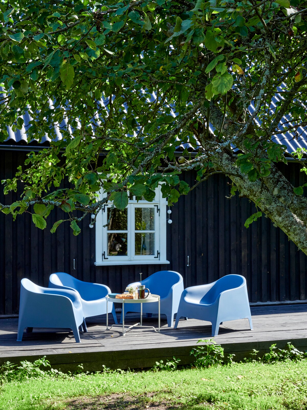 A seating area features four blue chairs arranged around a small table on a wooden platform. A tree provides shade overhead, and a white-framed window is visible in the background, framed by the dark wood of the house.