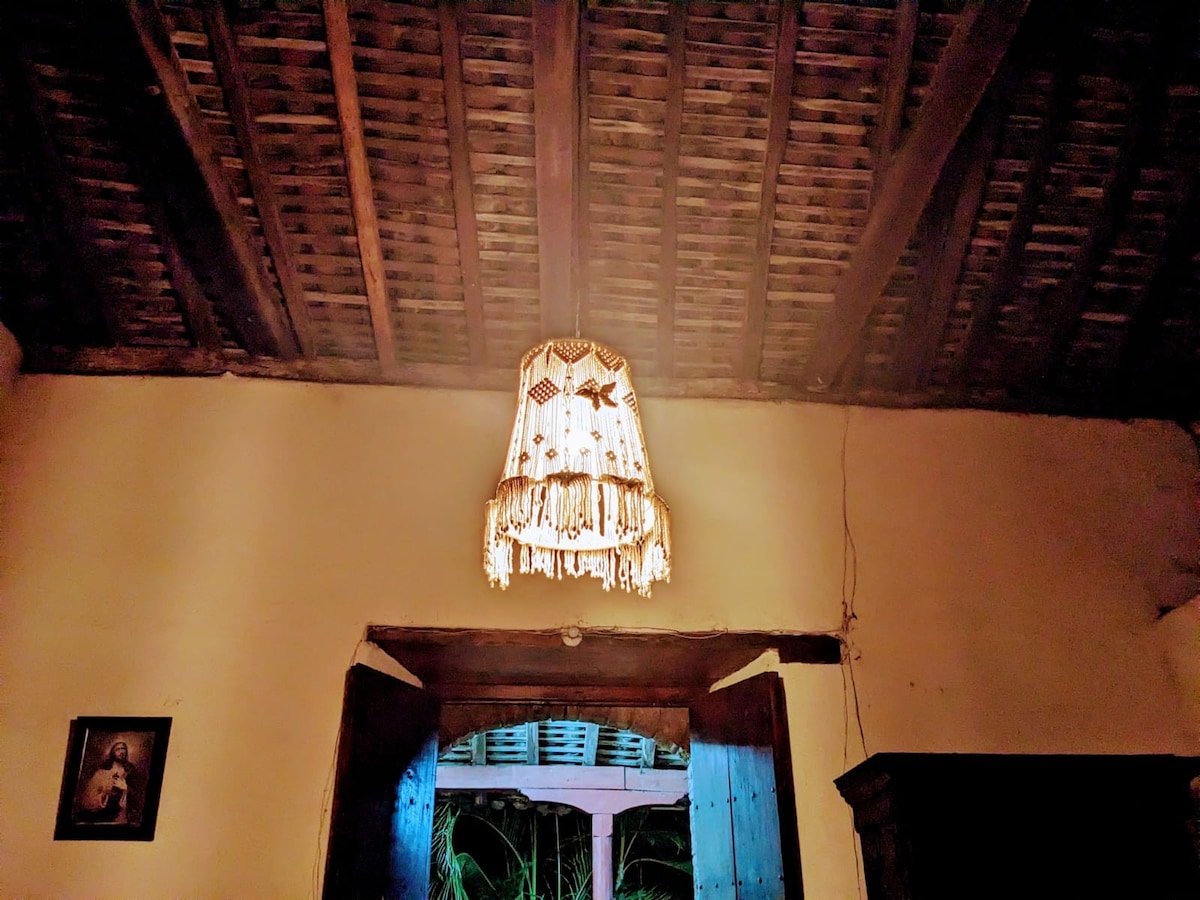 A handcrafted lamp hangs from a wooden ceiling, providing soft light that highlights the textured beams above. An open doorway reveals glimpses of outdoor greenery, while a framed artwork is positioned on the wall, adding character to the space.