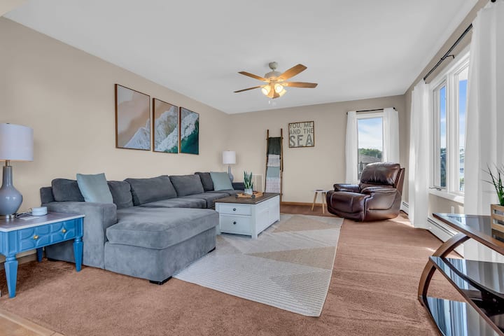 This cozy second living room offers a plush sectional, recliner, and large windows with beautiful lagoon views. Perfect for family movie nights or quiet relaxation, the space is both inviting and functional with charming coastal decor.