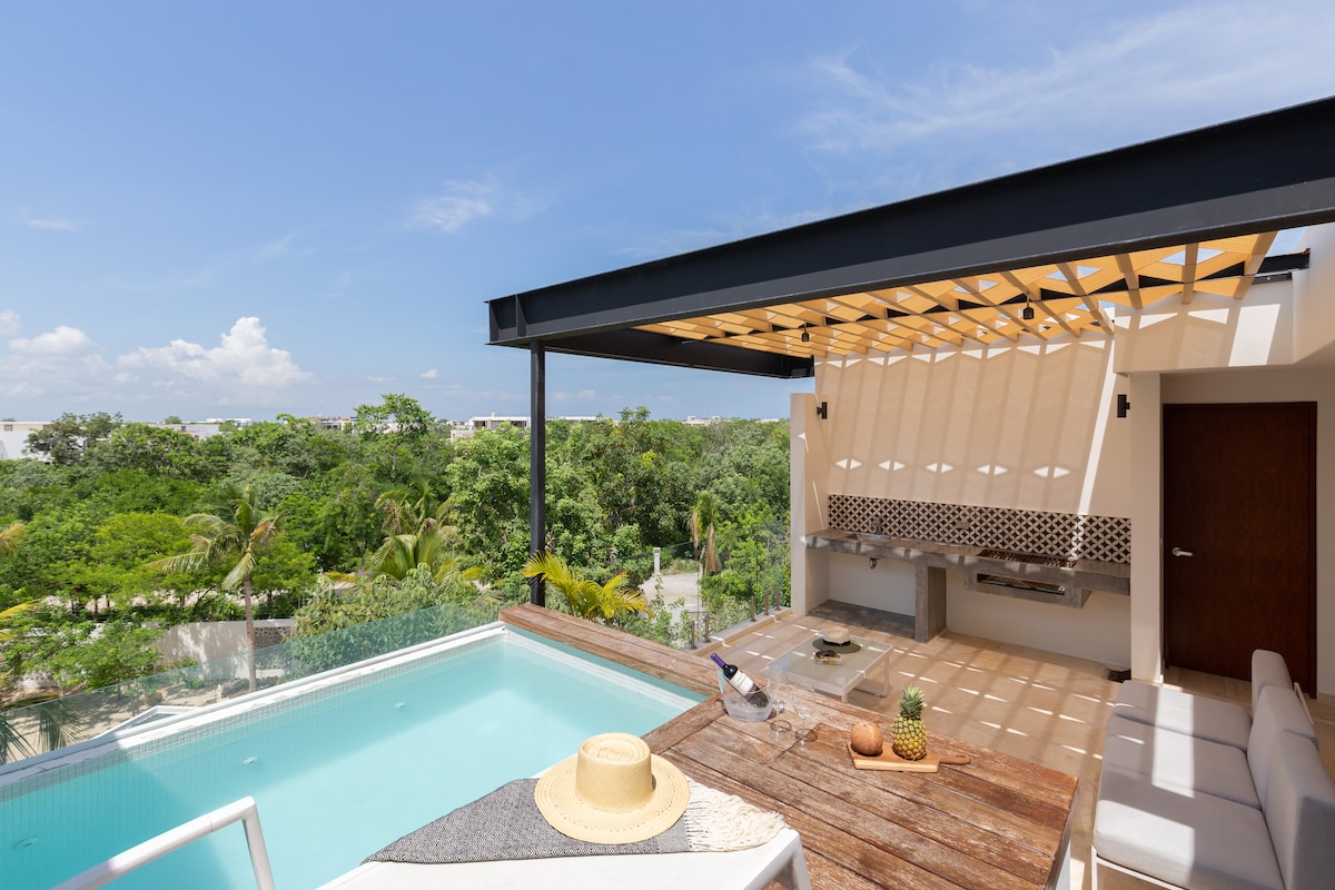 A private rooftop terrace overlooks lush greenery and features a plunge pool with clear blue water. A comfortable seating area is arranged next to the pool, with a shaded dining space that includes a built-in grill. A hat and decorative items are placed on the wooden table.