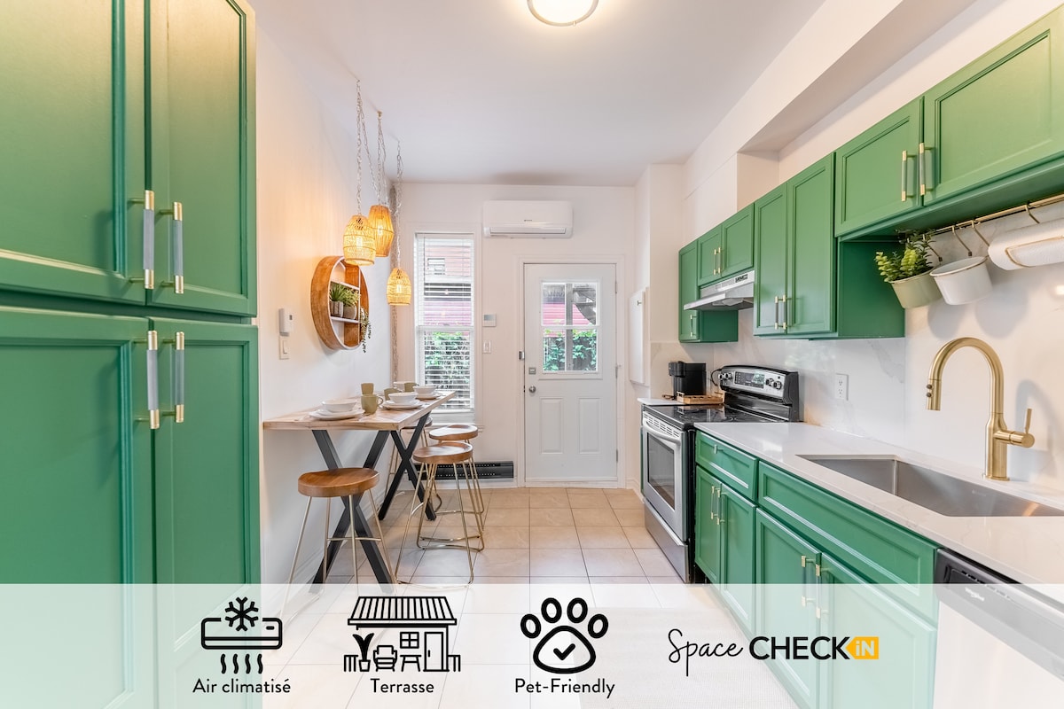 A modern kitchen features green cabinetry and stainless steel appliances. A small dining area with two bar stools is positioned next to a window. Soft lighting from pendant fixtures creates a warm ambiance, while a door leads to a private terrace.