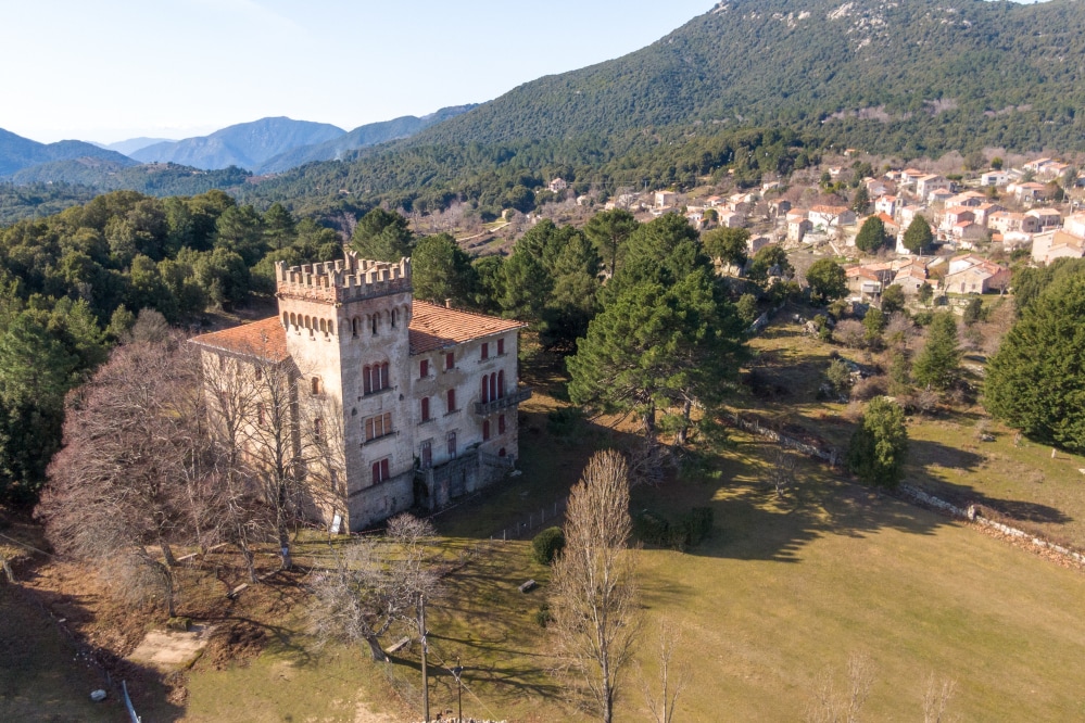 The image showcases a stately building with a medieval-style tower, set against a backdrop of rolling hills and a nearby village. The surrounding landscape features a mix of trees and open green spaces, providing a serene environment.