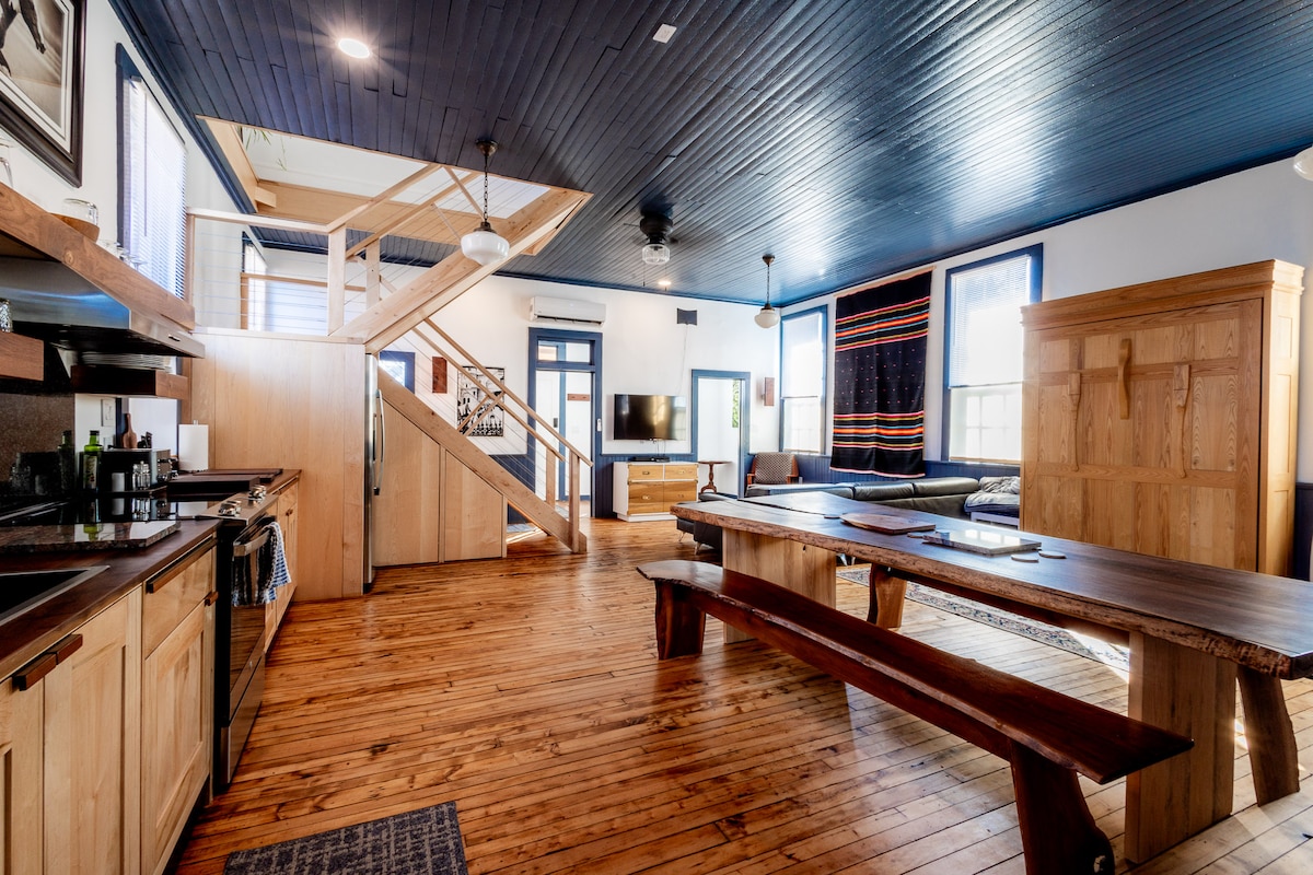 An open living area showcases warm wooden flooring and a high ceiling with a blue-painted surface. A full kitchen is equipped along one side, while a large dining table made of dark wood occupies the center. Comfortable seating is arranged, and a staircase leads to the upper level.