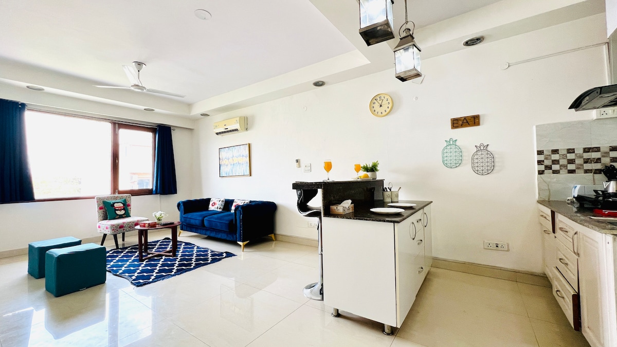 A bright and airy open-plan living area is presented, featuring a cozy blue couch, a dining space with bar stools, and an adequately equipped kitchen area. Large windows allow natural light to fill the room, complemented by cheerful wall decor and a decorative rug on the floor.