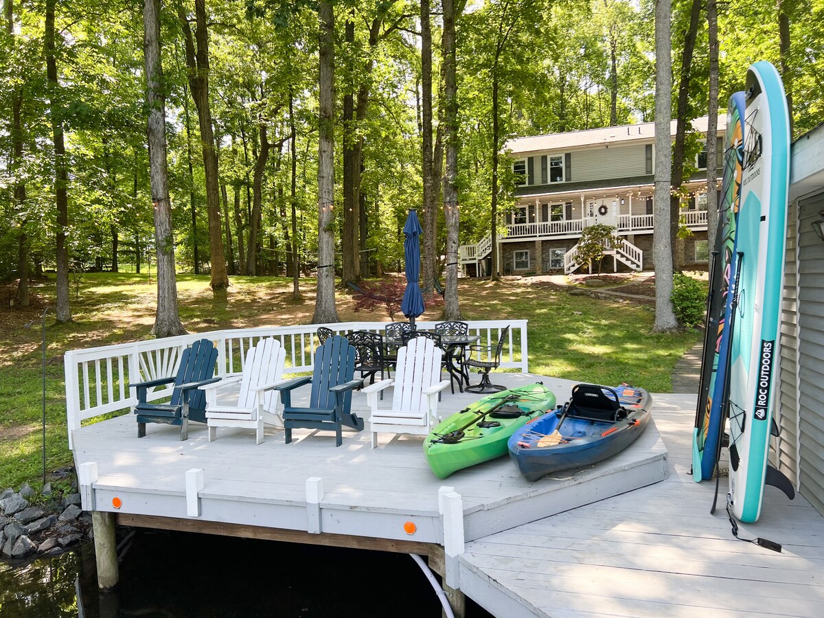 A spacious dock is outfitted with several adirondack chairs arranged for relaxation. Two kayaks in bright colors and a paddleboard are nestled alongside the dock. The serene setting is framed by lush trees, while the two-story home can be seen in the background.