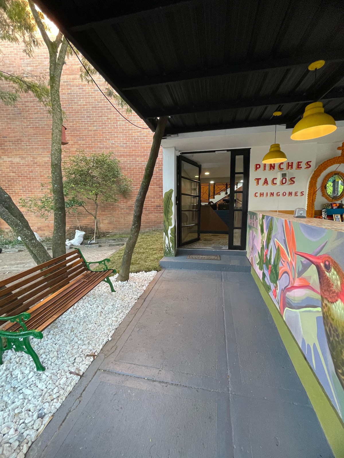 A welcoming entrance showcases a vibrant mural featuring a hummingbird. A bench made of wooden slats is positioned on a gravel pathway bordered by greenery. Large glass doors lead into the interior space, enhancing the inviting atmosphere.