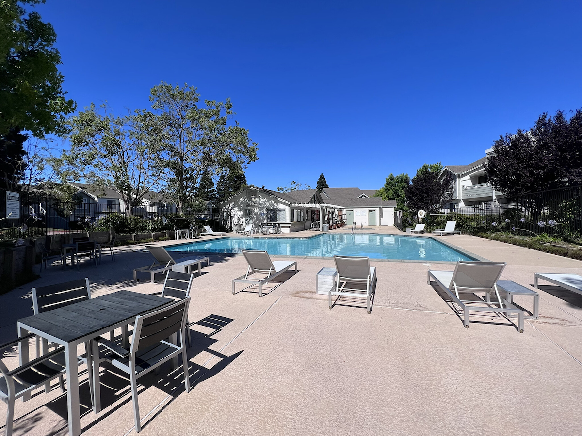 A tranquil pool area is surrounded by lounge chairs and dining tables. Lush greenery and well-maintained landscaping complement the space, while a clear blue sky adds to the inviting atmosphere of relaxation and leisure.