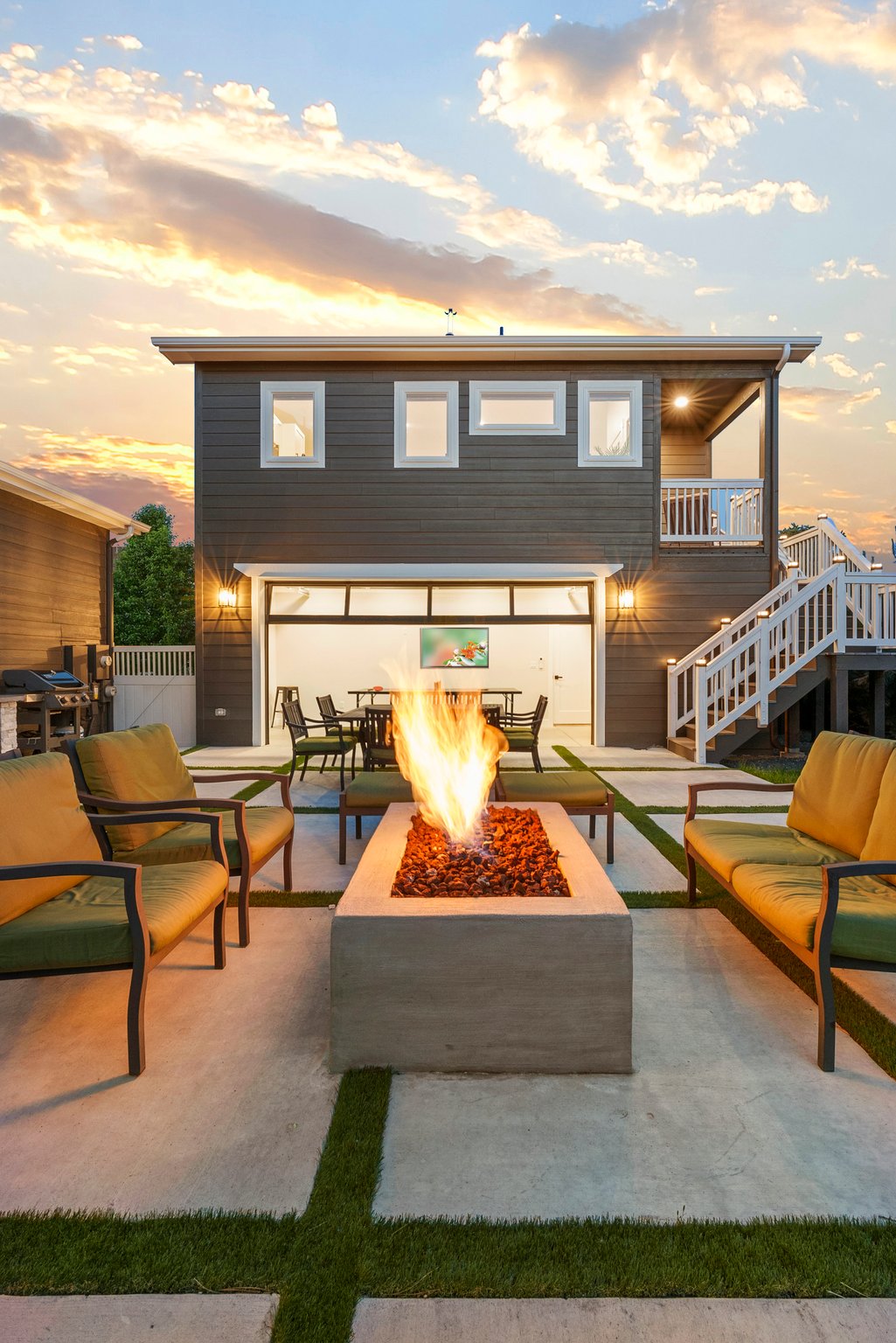 An outdoor patio area is furnished with green chairs surrounding a modern fire pit. The backdrop features a two-story building with large windows, and soft natural light illuminates the space as the sun sets in the distance.