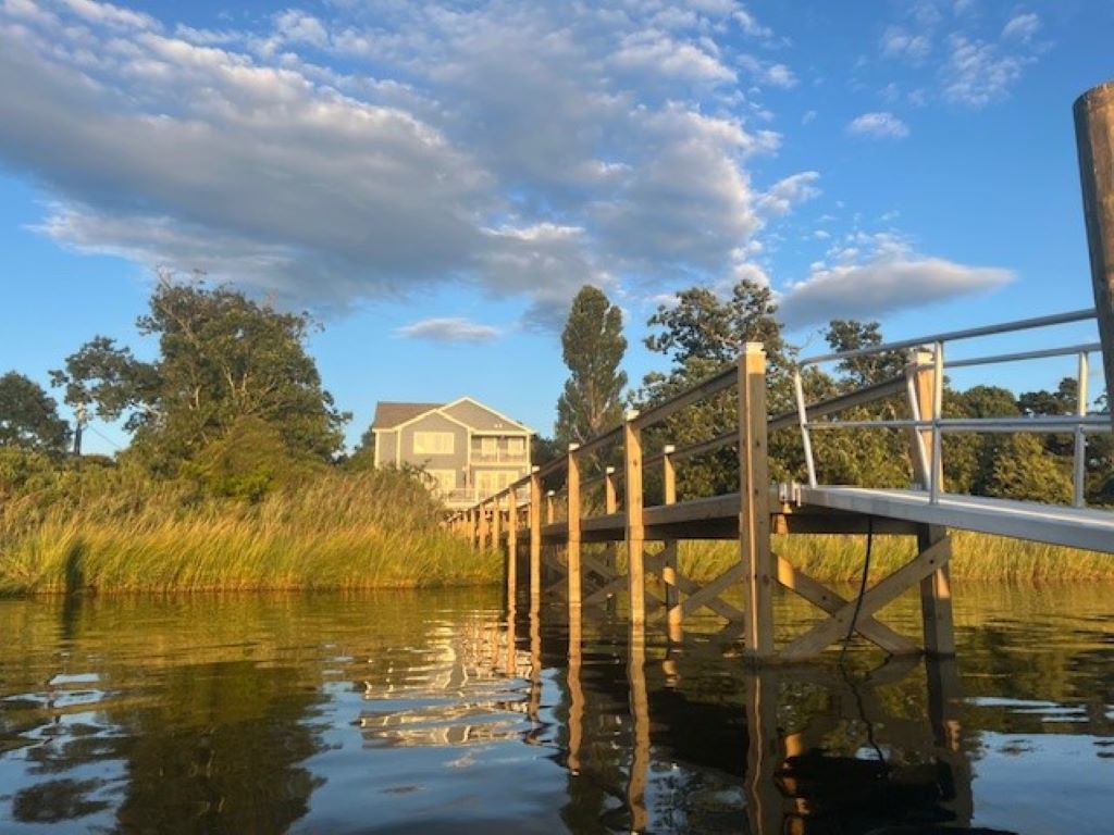 A private dock extends over the calm water, leading to a newly built waterfront house surrounded by greenery. The structure, visible in the background, features multiple levels, large windows, and is framed by tall trees under a bright blue sky with scattered clouds.