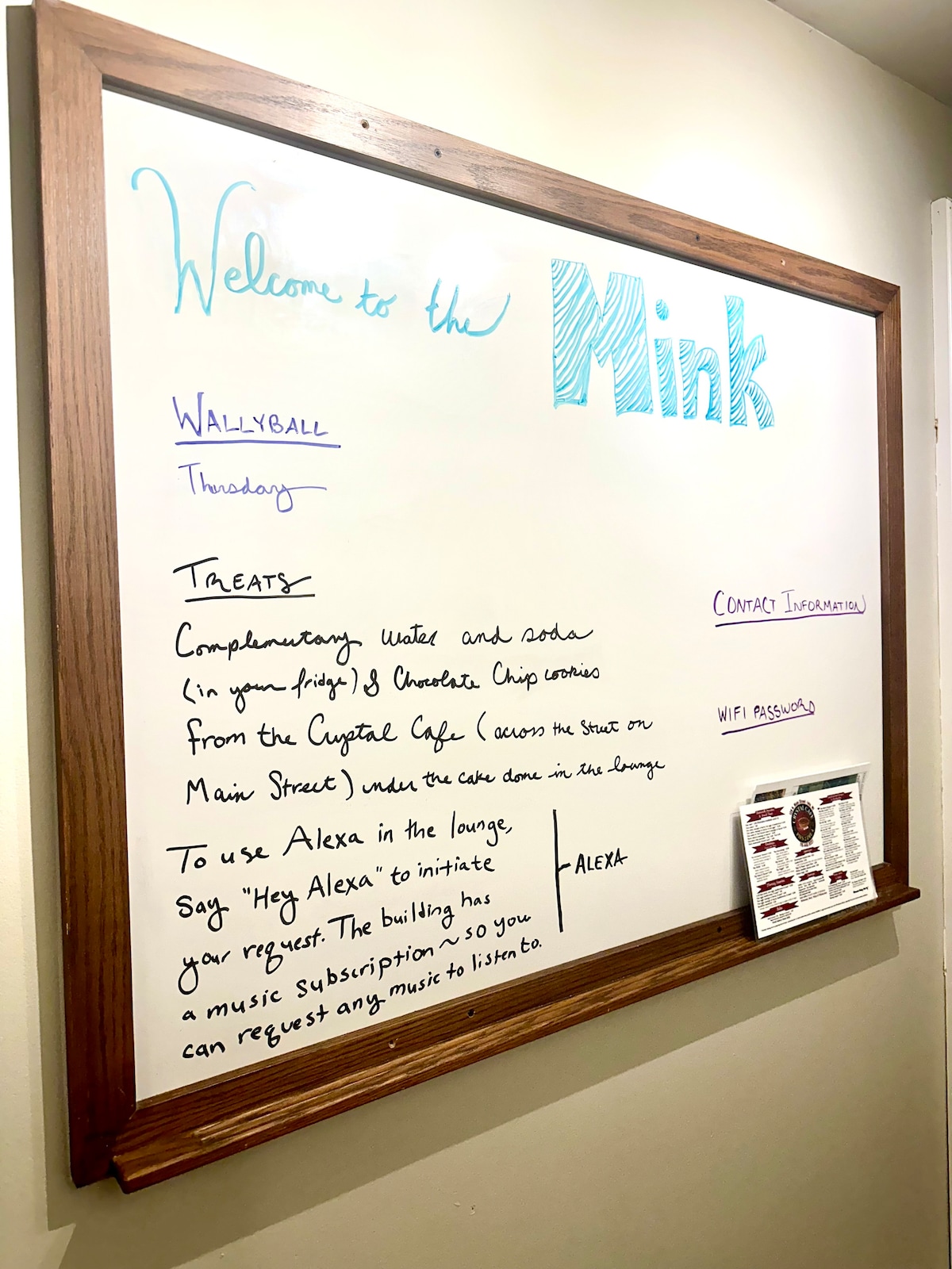 A large whiteboard in the hallway displays information and announcements. The top section welcomes guests to the Mink. Below, details about activities like wallyball and offerings of complimentary treats from nearby cafes are stated. Instructions for using the in-house Alexa device are also included.
