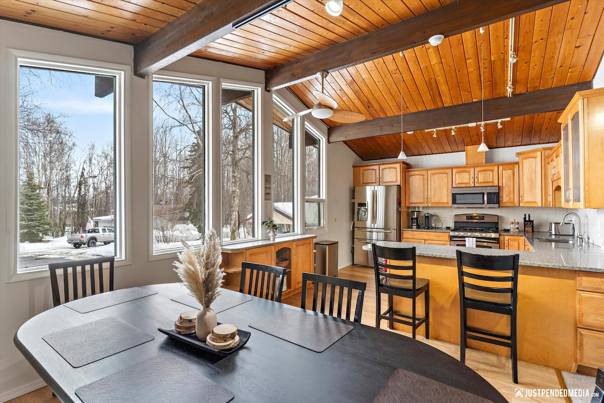An open kitchen and dining area features wooden cabinetry and modern appliances. Large windows allow natural light to fill the space, showcasing the outdoor view. A round dining table is set with place settings, and a breakfast bar with stools complements the layout.