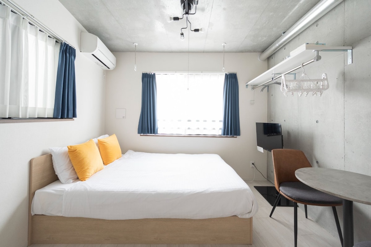 A spacious room is furnished with a comfortable bed featuring orange accent pillows. Natural light enters through a window dressed with blue curtains. An air conditioning unit is mounted on the wall, and a small dining table with a chair is positioned in the corner.