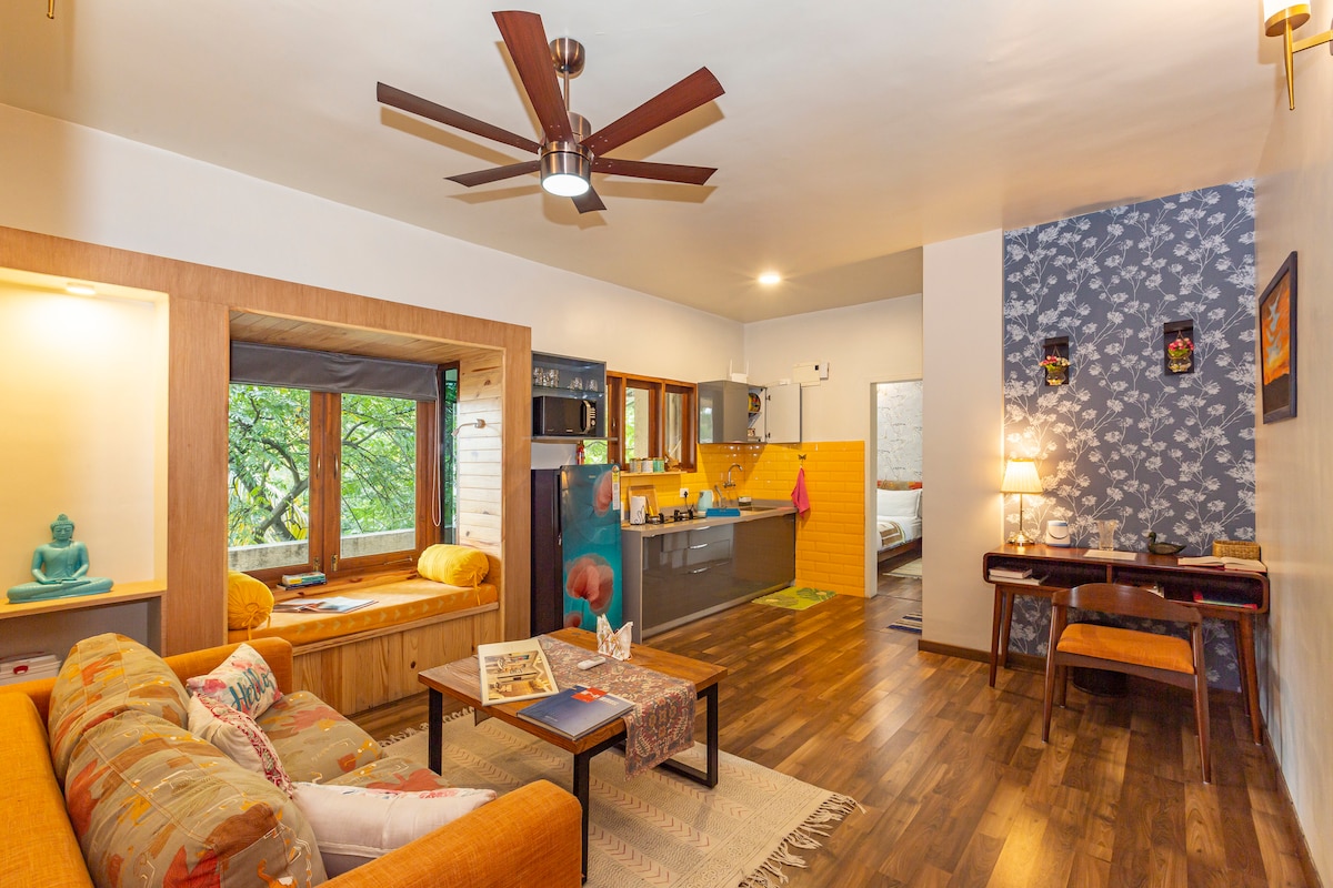 A cozy living area is featured, showcasing a comfortable sofa, a reading nook by the window, and a stylish dining table. The kitchen is visible, furnished with modern appliances. Soft lighting complements the warm wood tones and decorative elements on the walls.