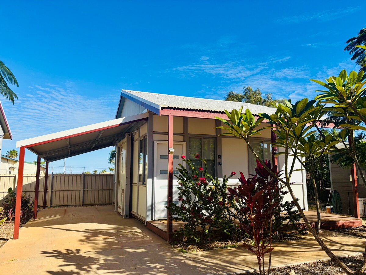 A single-story unit is viewed from the exterior, featuring a covered carport and a well-maintained garden. The vibrant green foliage complements the refreshing blue sky. The structure has a light-colored façade with a contrasting roof, providing a welcoming appearance.