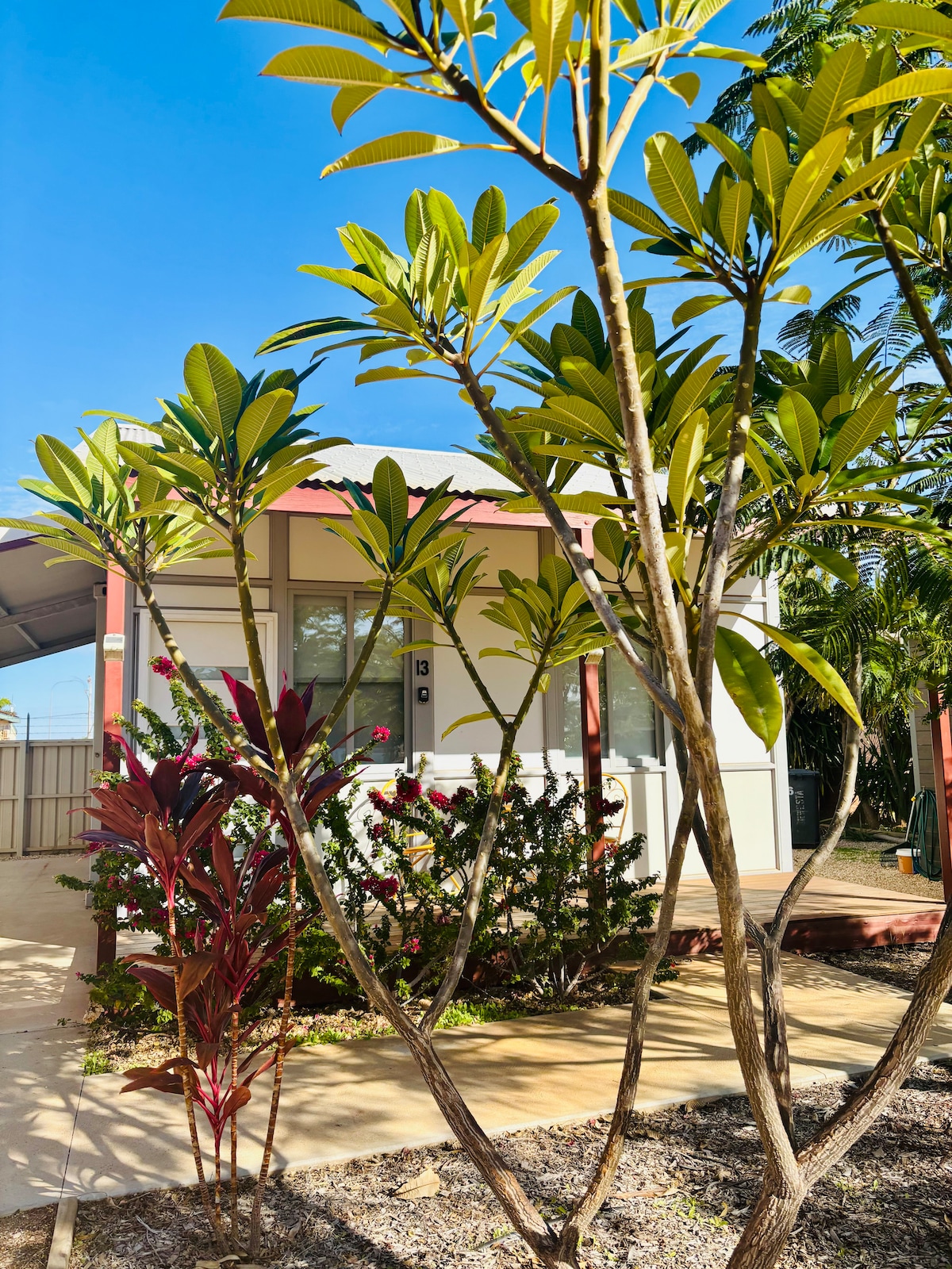 A charming exterior view of the unit is framed by lush greenery and colorful plants. The entrance features a traditional door and is surrounded by manicured landscaping, with a sunny blue sky visible above.