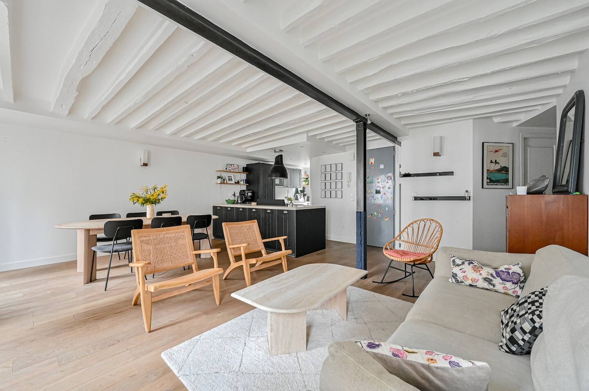 The open living area features a light wood floor and exposed ceiling beams. A spacious sofa is positioned near a low coffee table, while dining chairs surround a table in the background. The kitchen is visible, equipped with modern appliances and ample counter space.