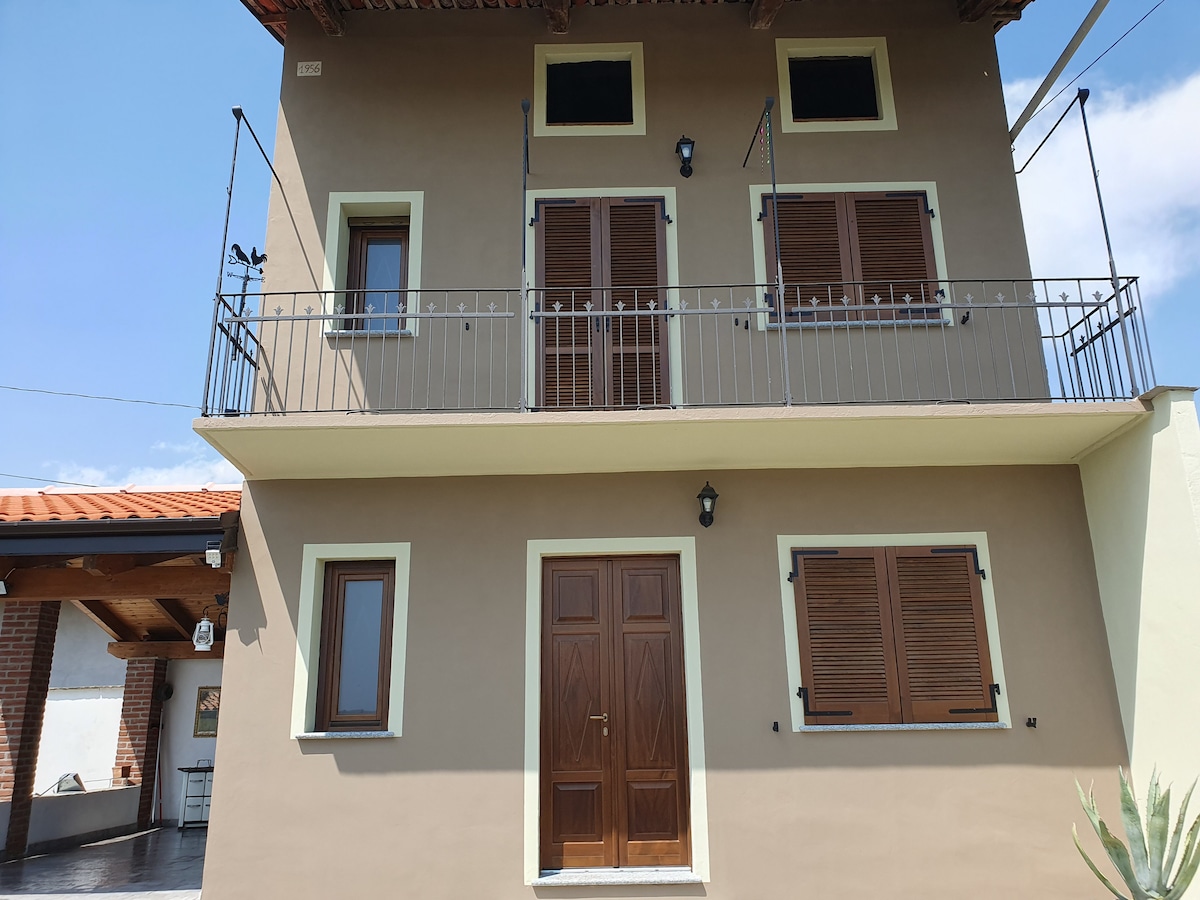 The two-story building features a symmetrical facade with wooden shutters on each window and a central double door entrance. Two balconies are situated above, providing open space. The exterior is painted in a neutral hue, complemented by a tile roof and clear blue sky.