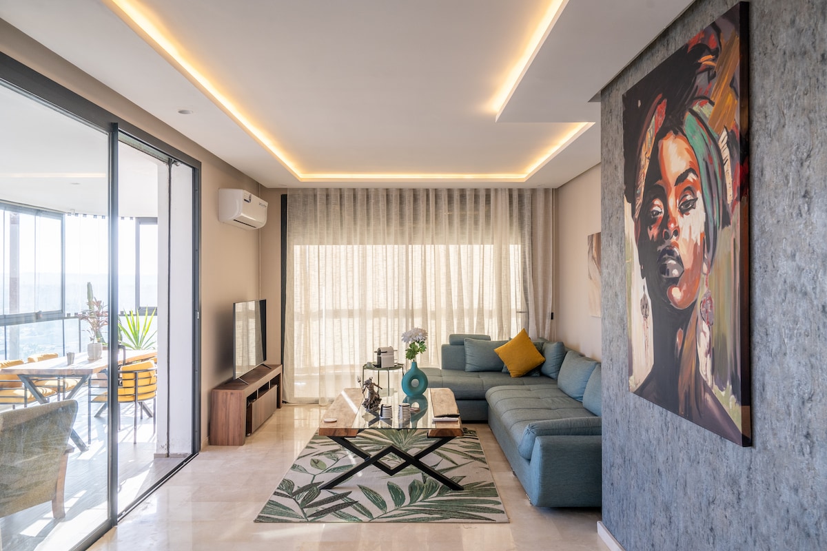 A modern living area features a spacious sectional sofa accented with colorful pillows. Natural light floods in through large glass doors, leading to a terrace. A contemporary piece of artwork adorns the wall, complementing the stylish decor and the patterned area rug beneath a glass coffee table.