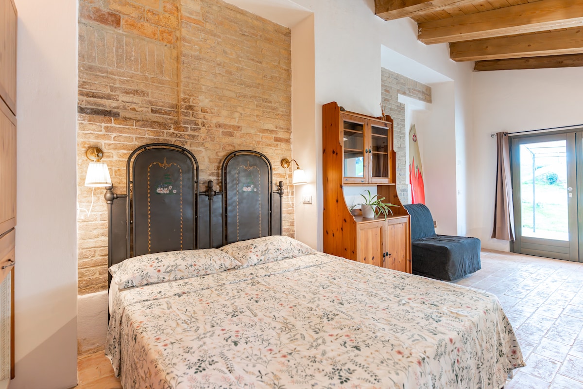 A spacious bedroom features a patterned floral bedspread over the double bed, complemented by two black metal headboards. Soft lighting is provided by sconces on either side. A wooden cabinet stands nearby, and a seating area is visible. Large glass doors lead to the garden.