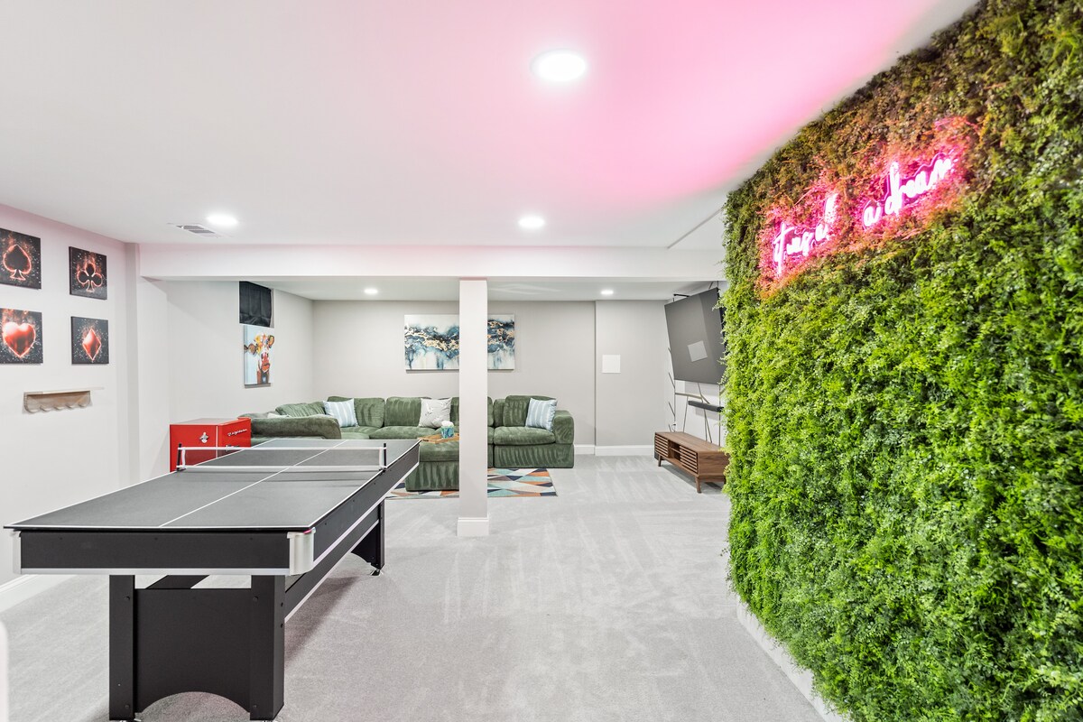 The basement recreation area includes a billiard and table tennis table, complemented by a comfortable seating area with a large sectional couch. Vibrant wall art is visible, alongside a green vertical garden feature and soft lighting that adds to the inviting ambiance.