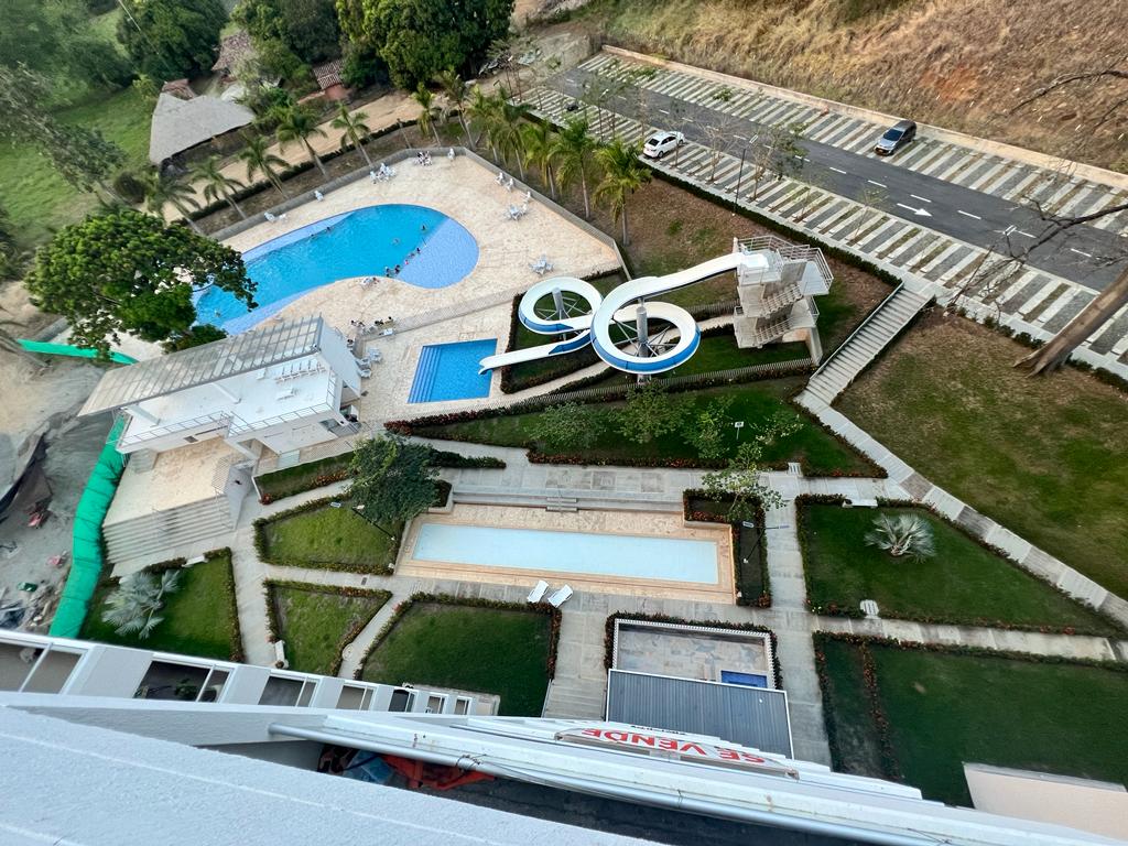 An aerial view of a resort-style recreational area reveals multiple swimming pools, one featuring a water slide. Surrounding green spaces are landscaped with well-defined paths, palm trees, and lounge areas, providing a relaxed atmosphere suitable for families.