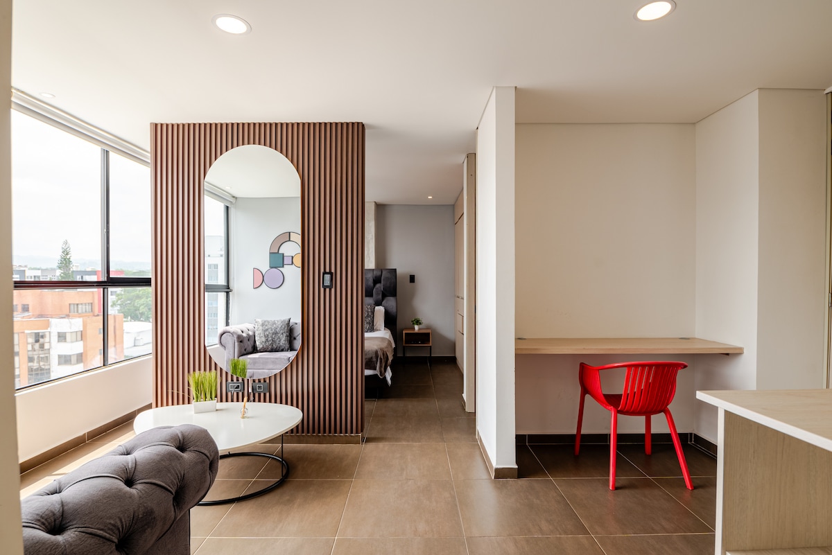 A well-lit living space features a comfortable gray sofa complemented by artistic decor. A round coffee table sits in front, alongside a red chair at a simple desk. Large windows allow natural light to fill the room, enhancing the modern finishes.
