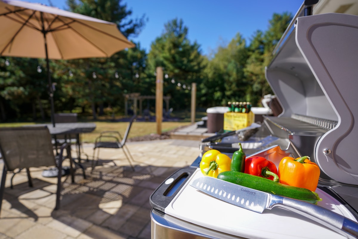 A barbecue grill sits prominently in the foreground, displaying fresh peppers and zucchini, ready for cooking. In the background, a patio umbrella shades a table with several chairs, surrounded by trees and a well-maintained lawn.