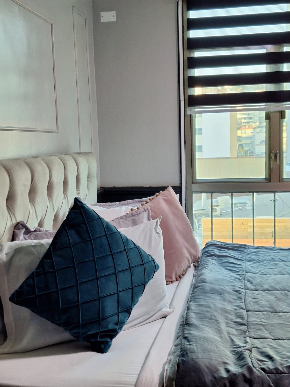 A well-appointed sleeping area features a spacious bed adorned with a combination of textured and solid-colored pillows. Natural light filters through partially closed blinds, enhancing the modern decor. A window provides a view of the bustling surroundings outside, creating a bright yet comfortable ambiance.