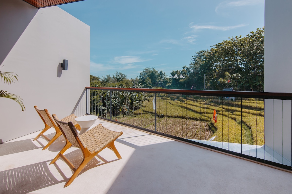 A balcony features two woven chairs and a small round table, offering a panoramic view of the lush rice paddies. Sunlight brightens the space, highlighting the natural surroundings and creating a peaceful outdoor area.