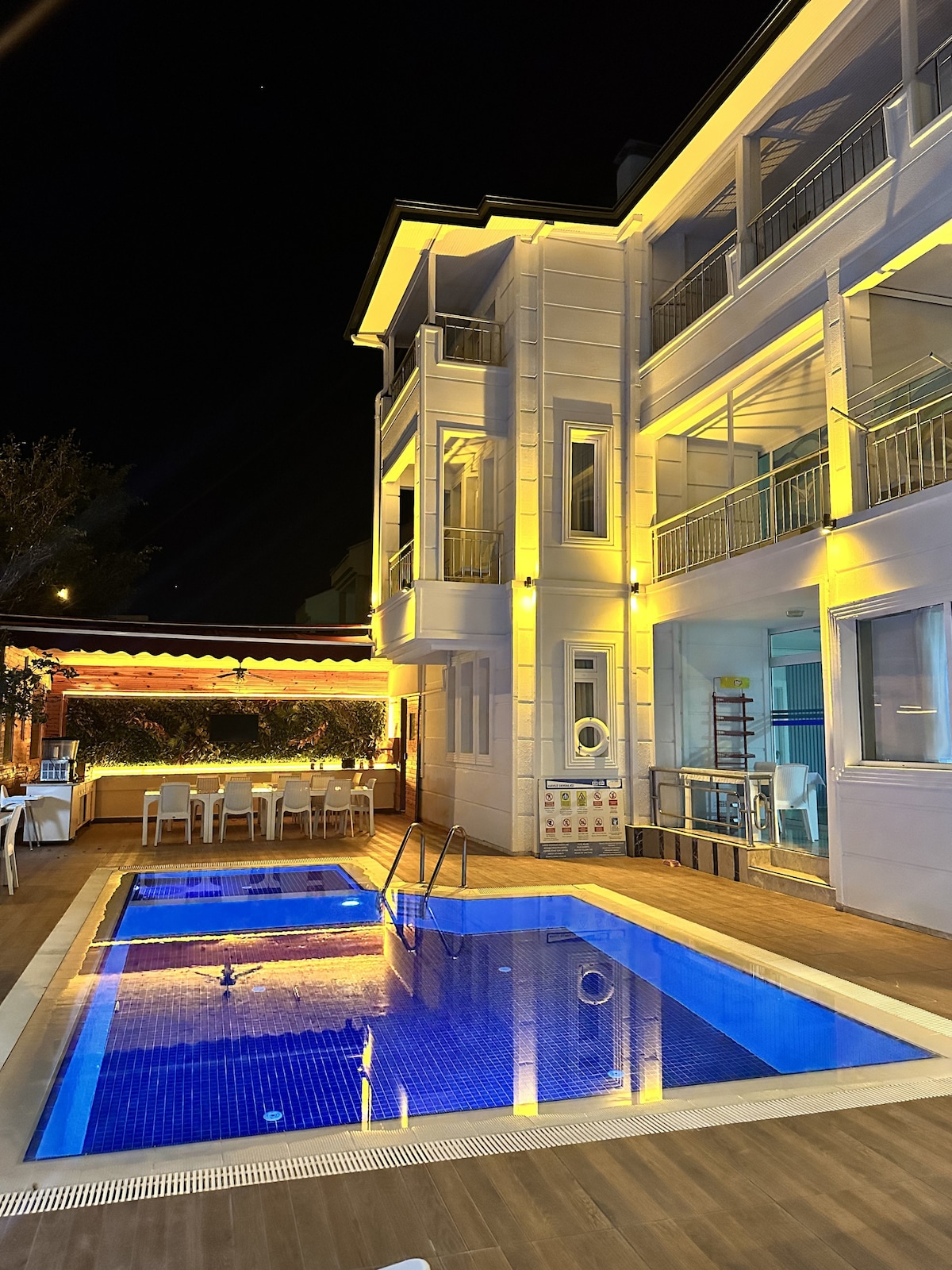 A charming outdoor area features a sparkling blue pool, illuminated by soft lighting. The multi-story building is adorned with elegant architectural details, and a dining space with white chairs can be seen beside the pool, providing a welcoming atmosphere for relaxation.