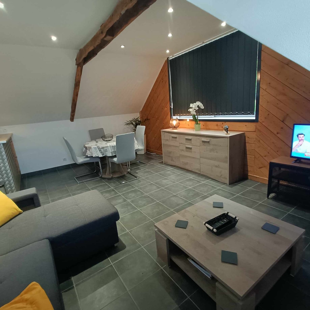 An open living space features a comfortable sofa and a small coffee table, accompanied by a dining table set for four. A sideboard is visible, and light filters in through large windows adorned with dark blinds, casting a warm ambiance across the room.