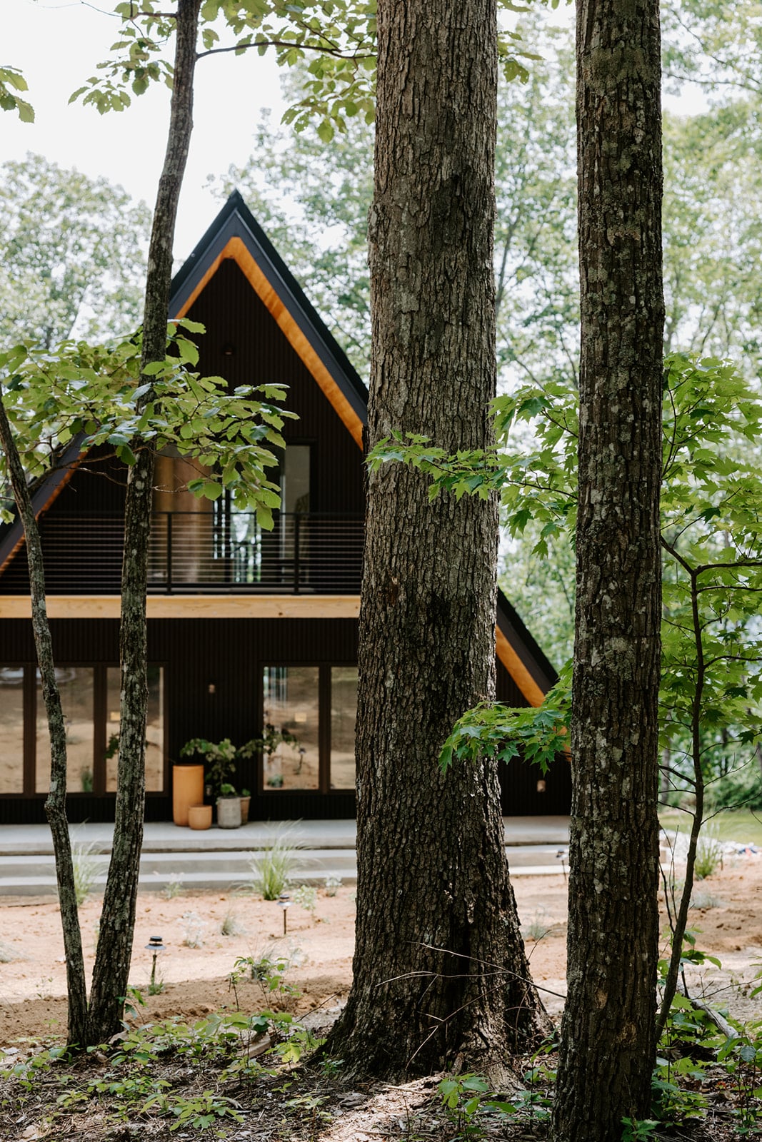 The A-frame structure is framed by tall trees, showcasing a modern design with large windows. A welcoming porch is visible, with potted plants placed beside the entrance. The natural surroundings highlight the secluded setting in the woods.